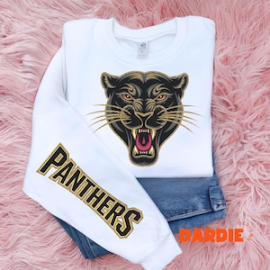 Pode incluir: Camisola branca com um gr&aacute;fico de pantera negra na frente e a palavra "PANTHERS" em glitter dourado na manga.