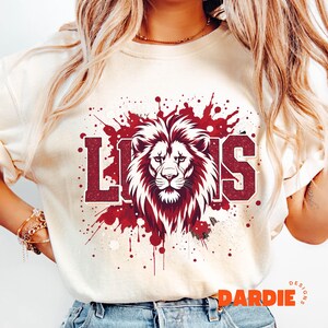 May include: Cream-colored t-shirt featuring a maroon lion graphic with the word "LIONS" in a collegiate font. The lion design is surrounded by maroon paint splatter details. The shirt has short sleeves and a crew neck.