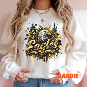 Peut inclure: Sweat-shirt blanc avec un motif d'aigle en paillettes dor&eacute;es et le mot "Eagles" en paillettes dor&eacute;es. Le motif a un effet de peinture &eacute;clabouss&eacute;e.