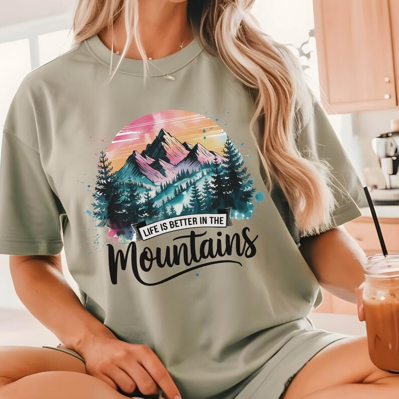 Op de afbeelding: Een lichtgroen T-shirt met een aquarel berglandschap en de tekst "Life is better in the Mountains".