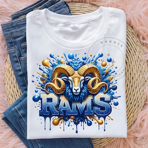 May include: White t-shirt featuring a graphic of a ram's head with large golden horns, blue fur, and the word "RAMS" in blue and white. The design includes blue and gold paint splashes. The shirt is displayed on top of blue jeans.