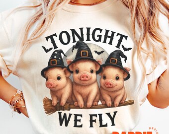 Könnte beinhalten: Cremefarbenes T-Shirt mit einem Halloween-Design. Es zeigt drei rosa Schweine mit schwarzen Hexenhüten, die auf einem Besen sitzen. Der Text "TONIGHT WE FLY" befindet sich über und unter den Schweinen, mit Fledermäusen und einem Mond im Hintergrund.