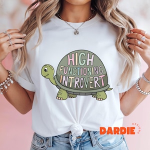 Könnte beinhalten: Weißes T-Shirt mit einer Comic-Schildkrötengrafik und dem Text "HIGH FUNCTIONING INTROVERT" in Regenbogenfarben.