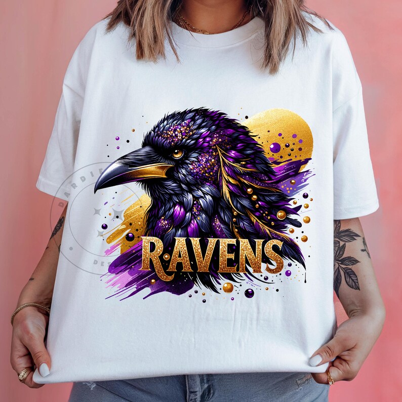 May include: White t-shirt featuring a detailed illustration of a raven's head. The raven is depicted with purple and gold feathers, a black beak, and the word "RAVENS" in gold lettering. The design includes gold accents and purple brushstrokes.