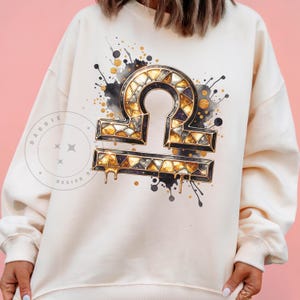 May include: A white sweatshirt with a gold and black Libra zodiac sign graphic. The sign is surrounded by black and gold watercolor splatters.