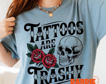 May include: A light blue t-shirt with a skull and roses graphic. The text on the shirt reads "Tattoos Are Trashy" with the word "DARDIE" in a smaller font below.