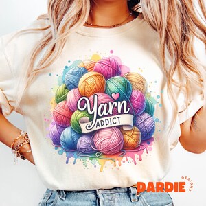 May include: Cream-colored t-shirt featuring a colorful graphic of yarn balls in various colors, including pink, blue, green, and yellow. The text "Yarn Addict" is displayed on a banner. Watercolor paint splatters surround the yarn balls.