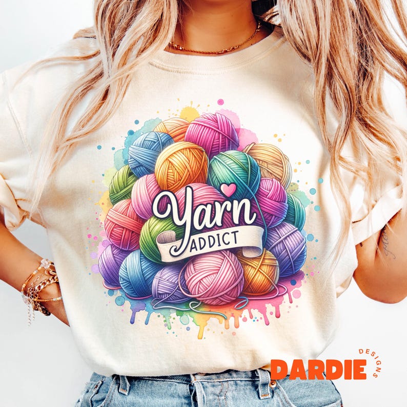 May include: Cream-colored t-shirt featuring a colorful graphic of yarn balls in various colors, including pink, blue, green, and yellow. The text "Yarn Addict" is displayed on a banner. Watercolor paint splatters surround the yarn balls.
