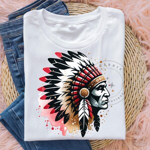 May include: White t-shirt with a graphic of a Native American man wearing a headdress with red, white, and black feathers. The headdress is decorated with gold glitter and red beads. The background is a watercolor wash of pink and red.