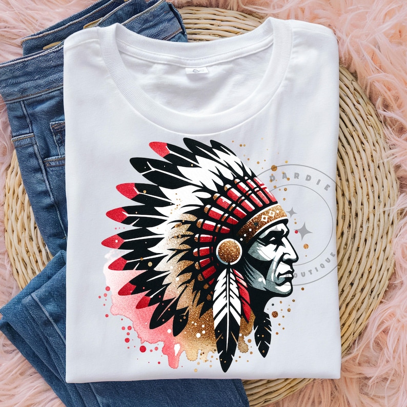 May include: White t-shirt with a graphic of a Native American man wearing a headdress with red, white, and black feathers. The headdress is decorated with gold glitter and red beads. The background is a watercolor wash of pink and red.