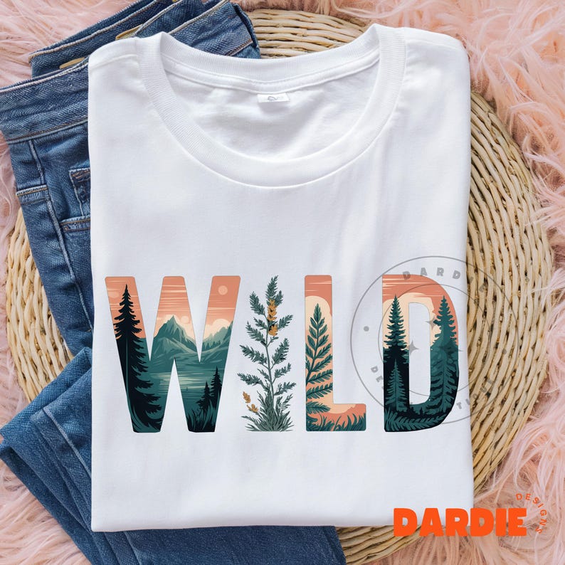 May include: White t-shirt with the word "WILD" in a nature-themed design. The letters are filled with images of mountains, trees, and foliage in shades of green, orange, and blue. The shirt is laid on top of blue jeans.