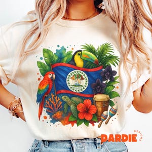 Puede incluir: Camiseta color crema con un diseño vibrante de la bandera de Belice con un tucán, un loro y flores tropicales. El diseño incluye una tortuga, un tambor y maracas, con la palabra "DARDIE" en naranja.