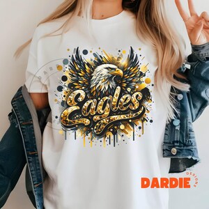 Peut inclure: T-shirt blanc avec un motif d'aigle en paillettes dor&eacute;es et le mot "Eagles" en paillettes dor&eacute;es. Le motif a un effet de peinture &eacute;clabouss&eacute;e noir et or.