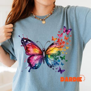 May include: A light blue t-shirt with a large watercolor butterfly design. The butterfly is made up of many colors, including pink, purple, blue, green, yellow, and orange. The butterfly is surrounded by smaller butterflies of various colors. The shirt has the text "DARDIE DESIGNS" in orange at the bottom.