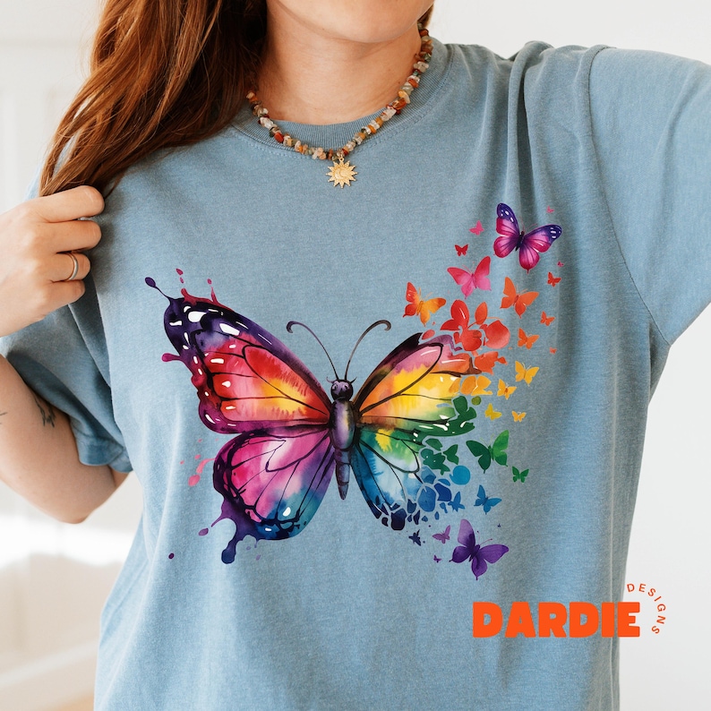 May include: A light blue t-shirt with a large watercolor butterfly design. The butterfly is made up of many colors, including pink, purple, blue, green, yellow, and orange. The butterfly is surrounded by smaller butterflies of various colors. The shirt has the text "DARDIE DESIGNS" in orange at the bottom.
