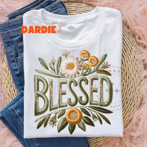 May include: White t-shirt with the word "BLESSED" embroidered in olive green, surrounded by floral embroidery in shades of cream, yellow, and green. The shirt is folded on top of blue jeans.