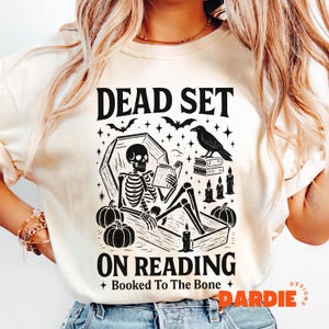 May include: Cream-colored t-shirt with a black graphic featuring a skeleton reading in a coffin, surrounded by pumpkins, candles, and a raven. The text reads "DEAD SET ON READING" and "Booked To The Bone."