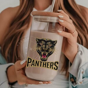 Pode incluir: Um copo de vidro transparente com um gr&aacute;fico de pantera negra e a palavra "PANTHERS" em glitter dourado. O copo tem uma tampa marrom e um canudo.