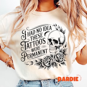 May include: Cream-colored t-shirt featuring a black skull graphic with roses and the text "I had no idea these tattoos were permanent." The design is a gothic style.