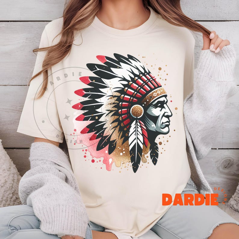 May include: A beige t-shirt with a graphic print of a Native American chief wearing a headdress with red, white, and black feathers. The graphic is surrounded by gold and pink watercolor splatters. The text "DARDIE" is printed in orange on the bottom right corner of the shirt.