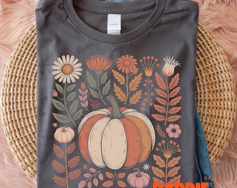 Op de afbeelding: Donkergrijs t-shirt met een herfstthema. De afbeelding bevat een pompoen, bloemen en bladeren in oranje, perzik en crèmekleurige tinten. Het shirt ligt op een geweven oppervlak met denim.