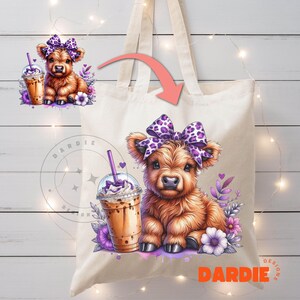 Pu&ograve; includere: Una borsa tote bianca con una mucca Highland marrone e bianca con un fiocco viola e leopardato. La mucca tiene un caff&egrave; freddo marrone e bianco. La borsa ha il testo "DARDIE DESIGNS" sul fondo.