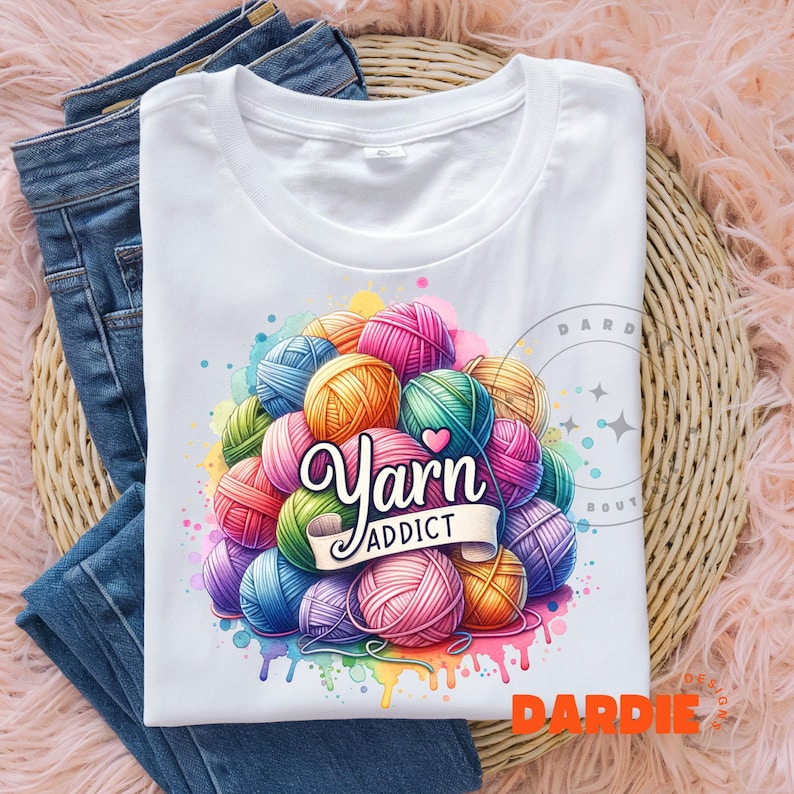 May include: White t-shirt featuring a colorful graphic of yarn balls in various colors, with the words "Yarn Addict" in a decorative font. The design includes a watercolor effect with rainbow paint drips. The shirt is folded on top of blue jeans.