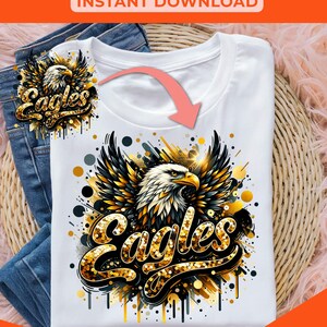 Peut inclure: Un t-shirt blanc avec un motif d'aigle en paillettes dor&eacute;es avec le mot "Eagles" en paillettes dor&eacute;es. Le motif est sur un t-shirt blanc avec un fond noir et or.