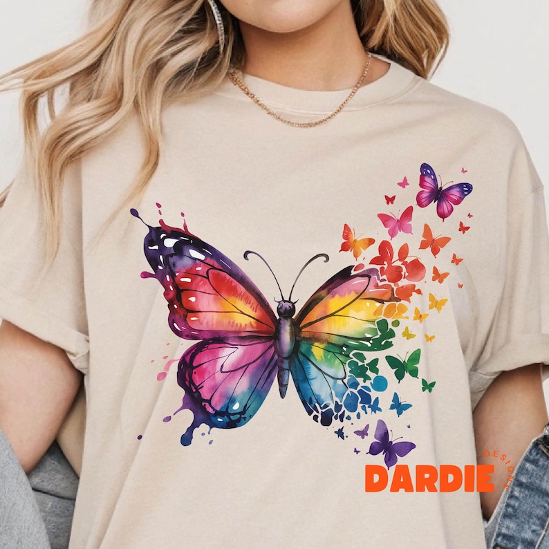 May include: A beige t-shirt with a large watercolor butterfly design in shades of pink, blue, yellow, and orange. The butterfly is surrounded by smaller butterflies in various colors.