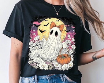 May include: Black t-shirt featuring a whimsical Halloween ghost design. The ghost, adorned with a bow, holds a pumpkin basket amidst flowers, bats, and a full moon. The artwork is set against a colorful sky.