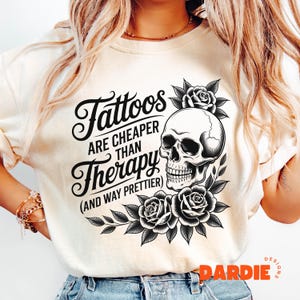 May include: Cream-colored sweatshirt featuring the text "Tattoos are cheaper than Therapy (and way prettier)" in black script. A detailed black and white skull graphic is surrounded by roses and leaves. The design is a bold statement.