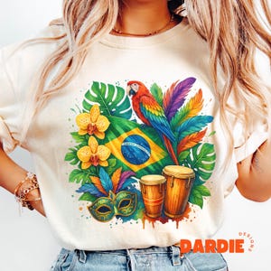May include: Cream-coloured t-shirt with a Brazilian-themed design. The graphic features a colourful parrot, the Brazilian flag, orchids, feathers, a carnival mask, and drums. The word "DARDIE" is printed at the bottom.