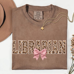 Può includere: Una maglietta marrone con la scritta "LIBRARIAN" in un motivo leopardato. Un fiocco rosa è sotto la parola e una collana a stella dorata è sulla maglietta. La maglietta è piegata su una superficie bianca.