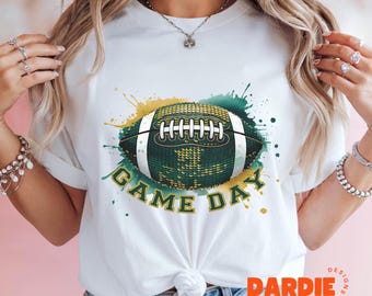 以下が含まれることがあります： 白地に緑と金のフットボールのグラフィックと「GAME DAY」の白い文字が書かれたTシャツ
