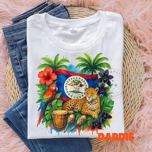 May include: White t-shirt featuring a colorful design with a flag, parrot, jaguar, and musical instruments. The design includes a red hibiscus flower, palm trees, and black flowers. The shirt is folded on top of blue jeans.