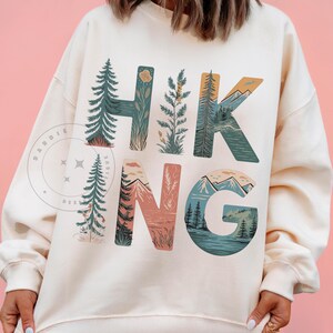Op de afbeelding: Een cr&egrave;mekleurig sweatshirt met het woord "HIKING" bedrukt in grote, kleurrijke letters. Elke letter is gevuld met een natuursc&egrave;ne, waaronder bergen, bomen en een meer.