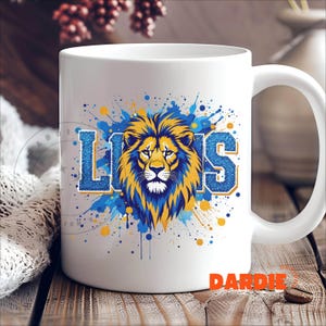 May include: White ceramic mug with a blue and yellow lion graphic and the word "LIONS" in blue glitter. The graphic has a watercolor effect with blue and yellow splatters.