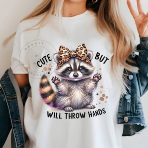 May include: White t-shirt with a cute raccoon wearing a leopard print bow. The text "Cute but will throw hands" is printed on the shirt.