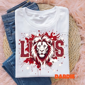 May include: White t-shirt with a red and white lion graphic and the word "LIONS" in red block letters. The design includes red paint splatters and is laid on top of blue jeans.
