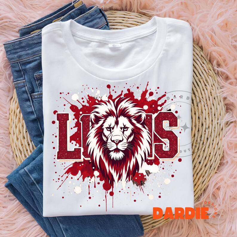 May include: White t-shirt with a red and white lion graphic and the word "LIONS" in red block letters. The design includes red paint splatters and is laid on top of blue jeans.
