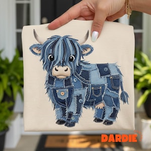 Könnte beinhalten: Cremefarbene Tragetasche mit einem Highland-Kuh-Design aus Jeansstoff-Flicken. Die Kuh hat blaue Jeansstoff-Flicken, Knöpfe und eine flauschige Jeansstoff-Mähne. Das Wort "DARDIE" ist in Orange unten rechts aufgedruckt.