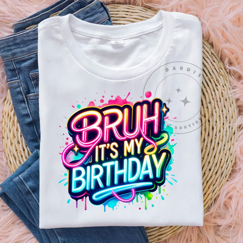 以下が含まれることがあります： 「Bruh it's my birthday」というネオン文字が書かれたカラフルなグラフィックデザインが施された白いTシャツです。