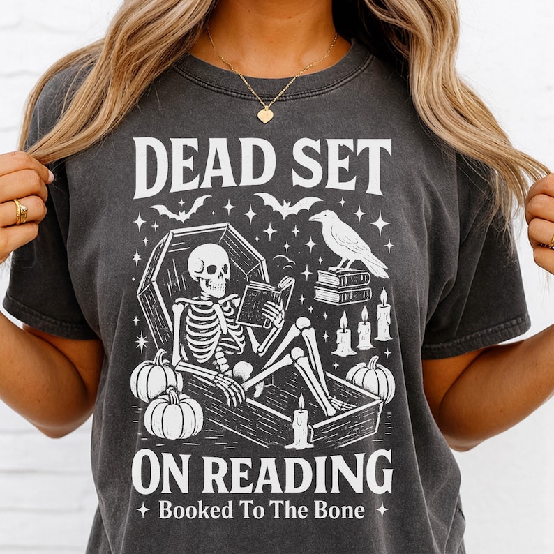 May include: Dark gray t-shirt with a Halloween-themed graphic. The design features a skeleton reading in a coffin, pumpkins, bats, and the text "DEAD SET ON READING" and "Booked To The Bone".