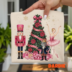 May include: A beige Christmas-themed decorative square featuring a pink nutcracker, a decorated Christmas tree, a ballerina, and a mouse king. The tree is adorned with red poinsettias and a pink ribbon. Presents are at the base of the tree.