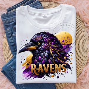 May include: White t-shirt featuring a detailed illustration of a raven's head with purple, gold, and black accents. The word "RAVENS" is displayed in gold lettering. The shirt is folded on top of blue jeans.