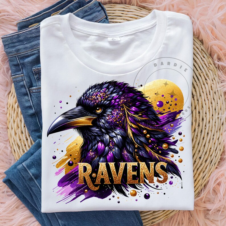 May include: White t-shirt featuring a detailed illustration of a raven's head with purple, gold, and black accents. The word "RAVENS" is displayed in gold lettering. The shirt is folded on top of blue jeans.