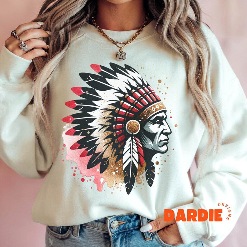 May include: A white sweatshirt with a colorful graphic of a Native American chief wearing a headdress. The headdress is decorated with red, white, and black feathers. The background of the graphic is a watercolor-style design with pink, gold, and black splatters. The text "DARDIE DESIGNS" is printed in orange on the bottom right corner of the sweatshirt.