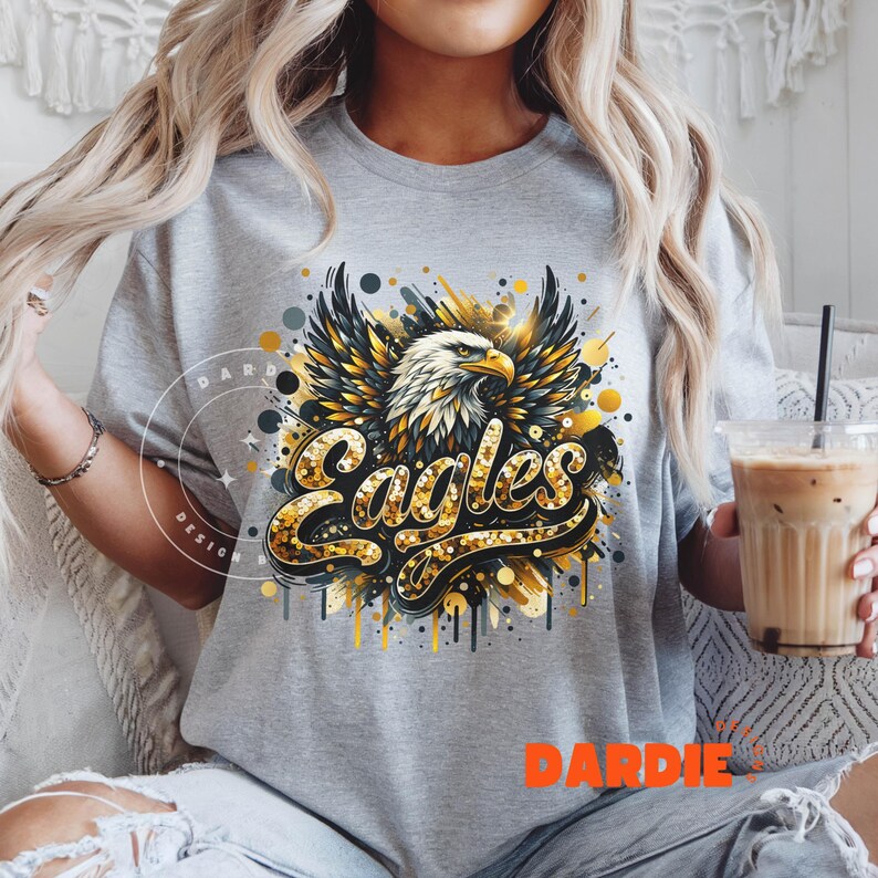 Peut inclure: Un t-shirt gris avec un motif d'aigle en paillettes dor&eacute;es et le mot "Eagles" en paillettes dor&eacute;es. Le motif a un effet de peinture &eacute;clabouss&eacute;e.