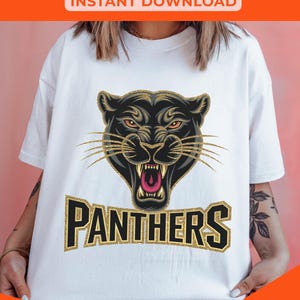 Pode incluir: Camiseta branca com um gr&aacute;fico de pantera negra e texto em glitter dourado que diz "PANTHERS".