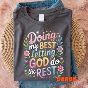 Pode incluir: T-shirt cinzenta escura com o texto "Doing my BEST Letting GOD do the REST" em letras florais coloridas. A camisa está dobrada sobre jeans e uma cesta de vime.